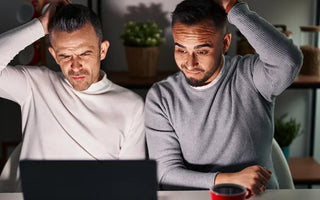 Two men are sitting at a table, looking confused at a laptop screen, struggling with finding poppers online. Both have one hand on their heads and are wearing casual long-sleeve shirts. In the background, there's a red mug and shelves adorned with plants.