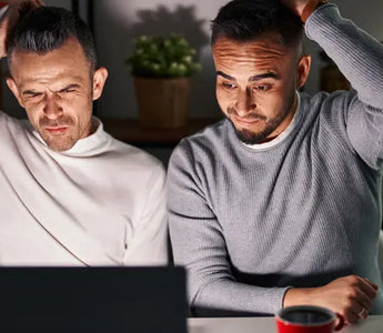 Two men are sitting at a table, looking confused at a laptop screen, struggling with finding poppers online. Both have one hand on their heads and are wearing casual long-sleeve shirts. In the background, there's a red mug and shelves adorned with plants.