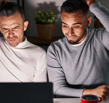 Two men are sitting at a table, looking confused at a laptop screen, struggling with finding poppers online. Both have one hand on their heads and are wearing casual long-sleeve shirts. In the background, there's a red mug and shelves adorned with plants.